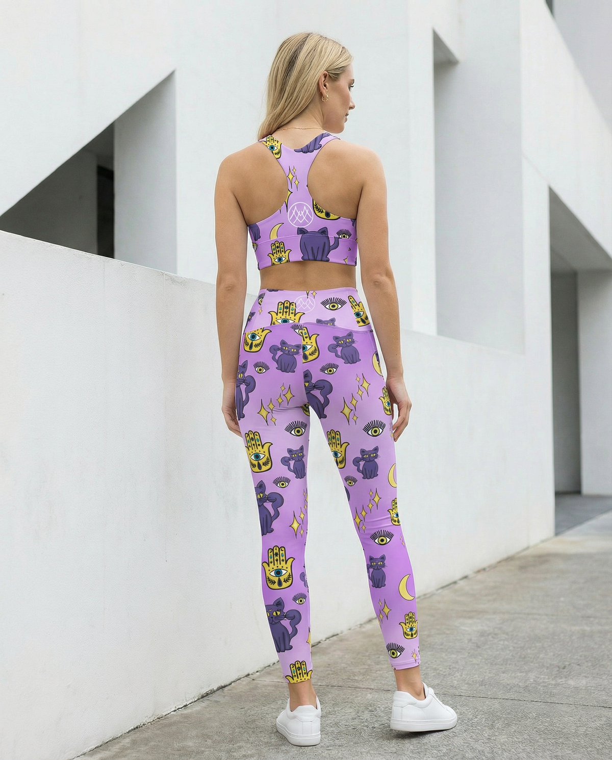 Woman wearing outfit of purple longline sports bra and purple leggings with purple cats and yellow evil eye pattern on them with side pockets Back view