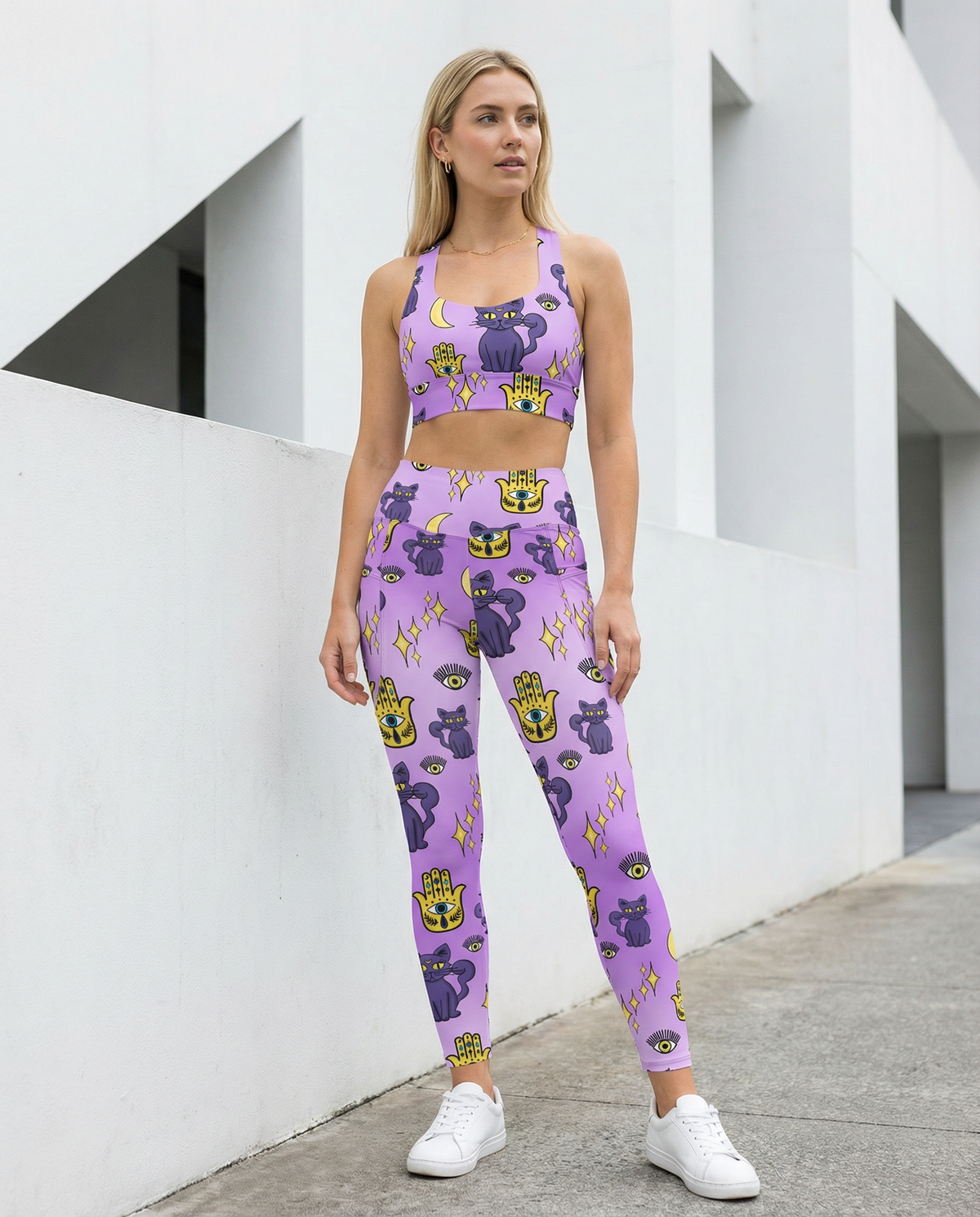 Woman wearing outfit of purple longline sports bra and purple leggings with purple cats and yellow evil eye pattern on them with side pockets front view
