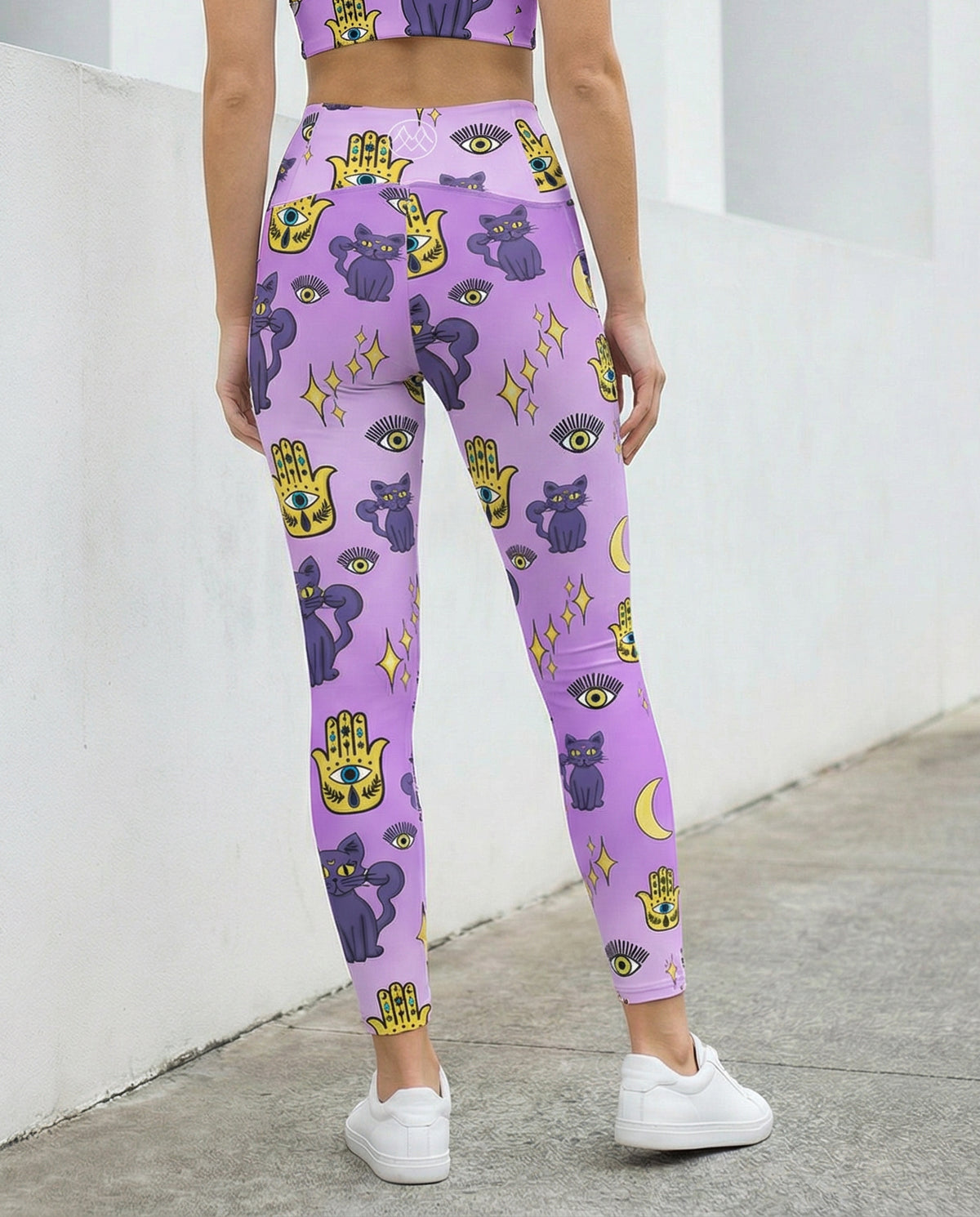 Woman wearing purple leggings with purple cats and yellow evil eye pattern on them with side pockets Back view